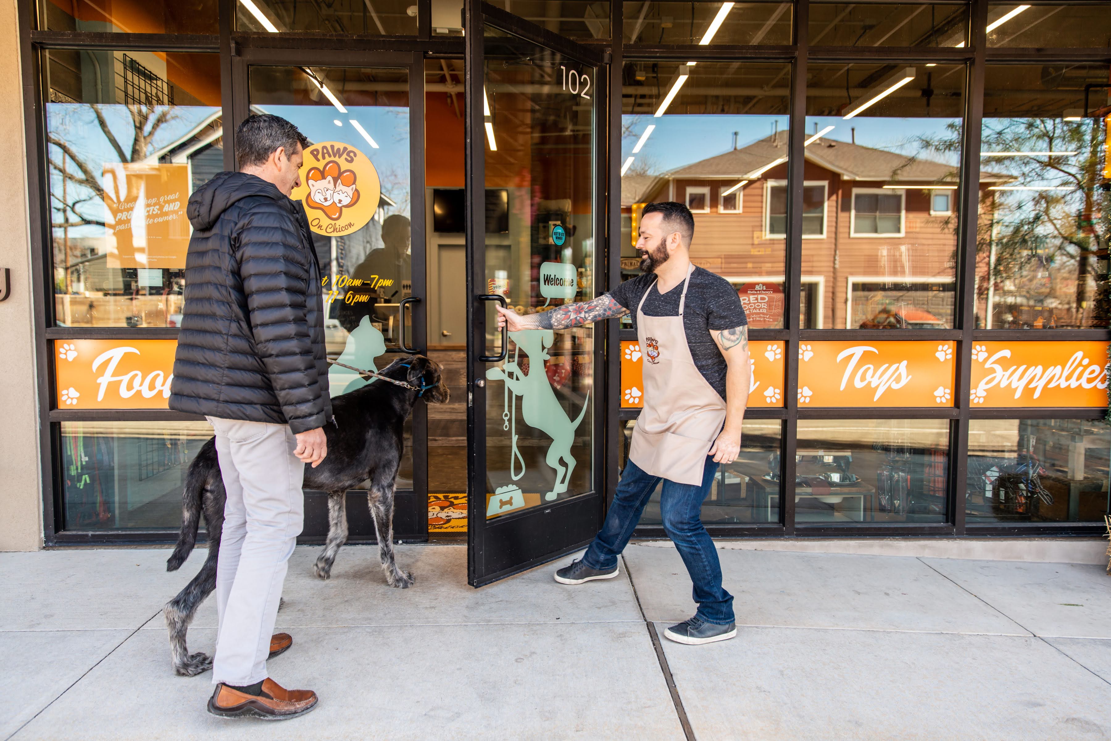 A man holds open the door to Paws on Chicon, an independent pet store, for another man who is walking a large black dog.